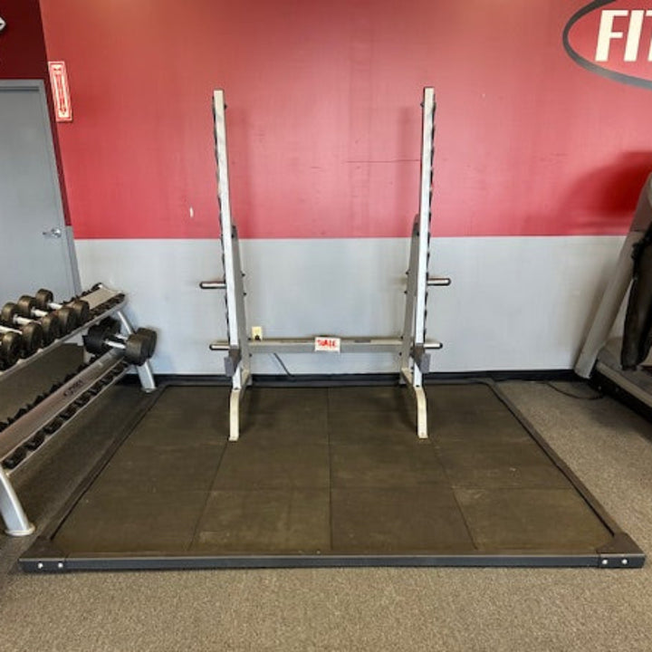 A white metal power rack with a barbell setup, positioned on a black rubber gym floor mat in a gym setting with a red and white wall background.