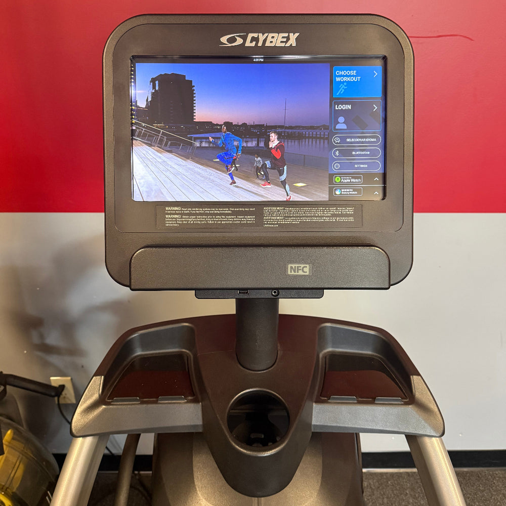 A Cybex treadmill shown from the front, featuring a large digital display screen on the console and a dark gray and silver frame, set against a red and white wall background.