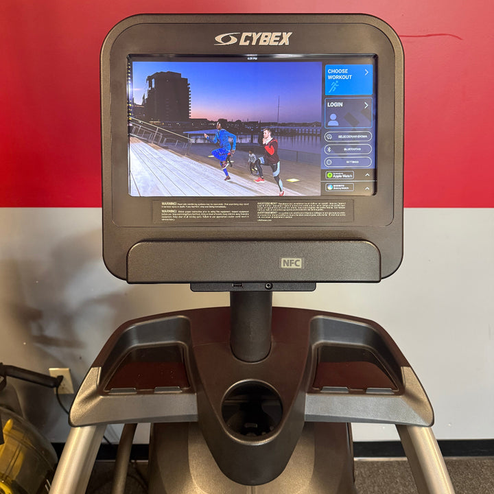 A Cybex treadmill shown from the front, featuring a large digital display screen on the console and a dark gray and silver frame, set against a red and white wall background.