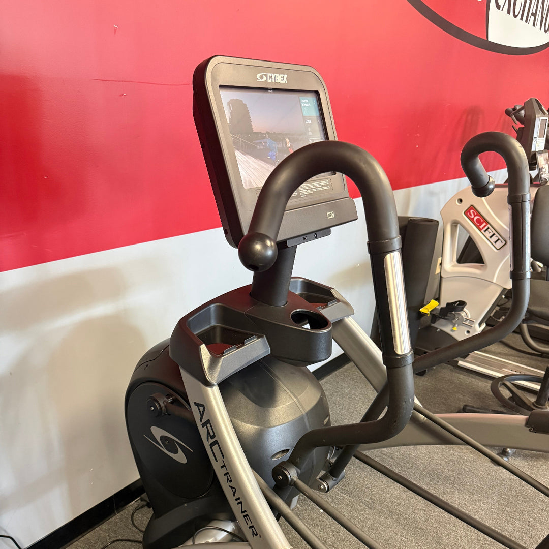 An Arc Trainer elliptical trainer with a digital display screen, set in a gym environment against a red and white wall.