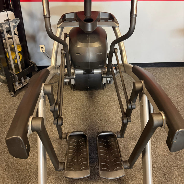 A Cybex elliptical trainer with a digital display screen, dark brown padded arms, and silver metal frame, photographed in a gym setting with a red and white wall background.