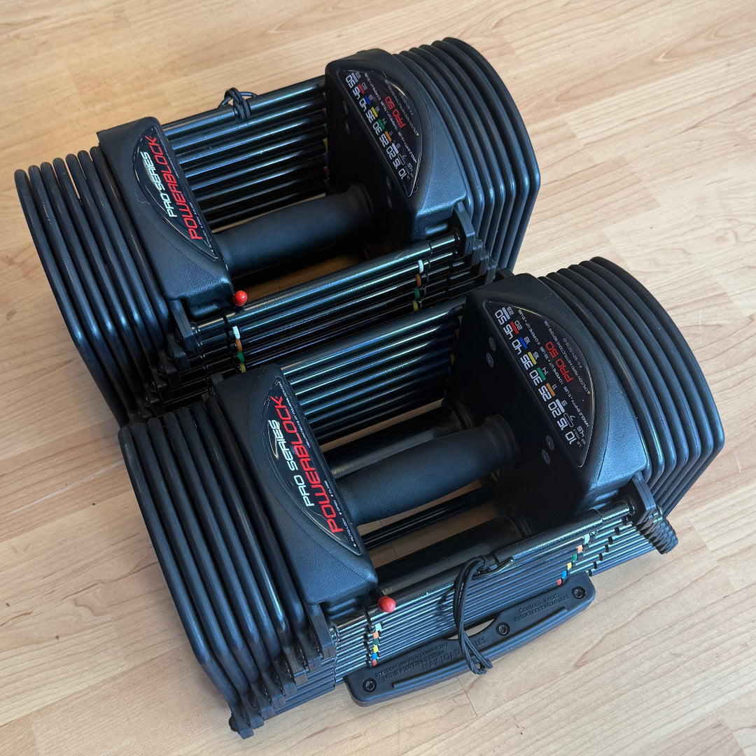 Two black adjustable dumbbells with ribbed handles and weight sliders, placed on a light wood floor. The front dumbbell shows the brand name &