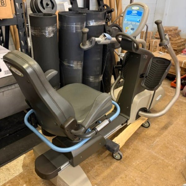 A home exercise machine with a gray seat, beige frame, and blue handlebar loop, photographed in a warehouse or storage area with exercise equipment in the background.