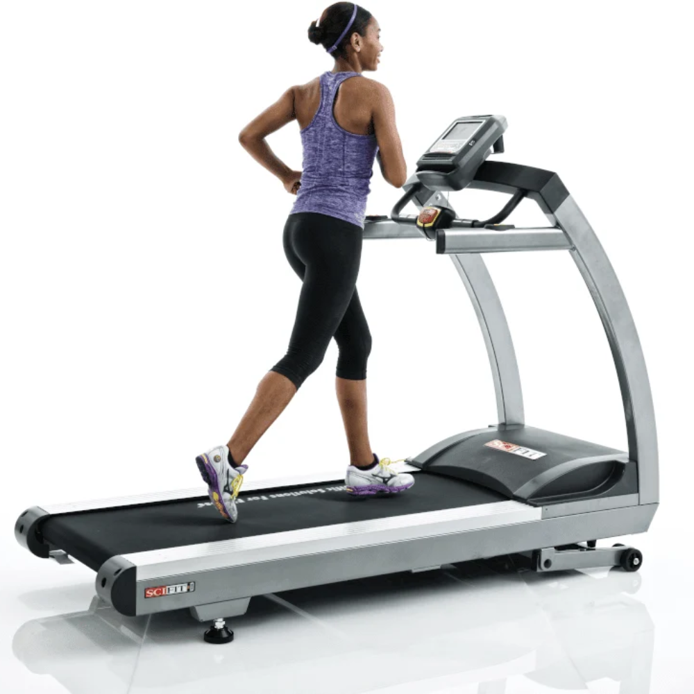 A woman running on a sleek DEKTR treadmill with a digital display screen, shown from a rear three-quarter angle against a clean white background.