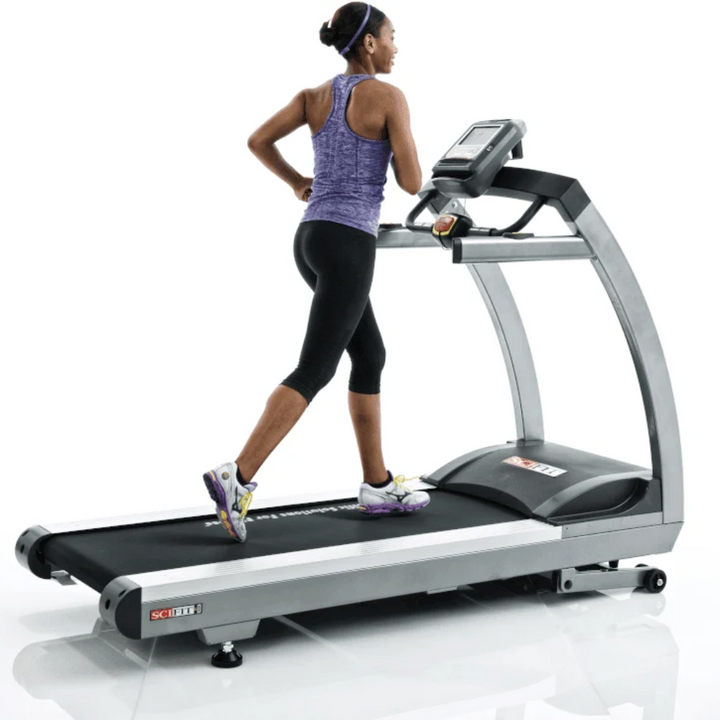 A woman running on a sleek DEKTR treadmill with a digital display screen, shown from a rear three-quarter angle against a clean white background.