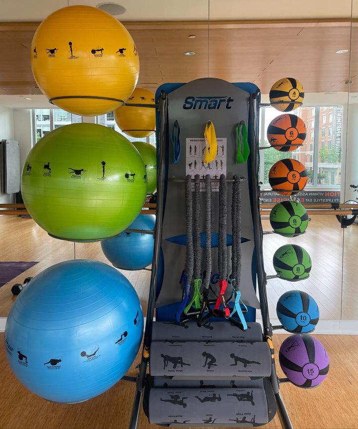 Gym equipment including colorful exercise balls and a storage unit labeled 'Smart' in a room with large windows.