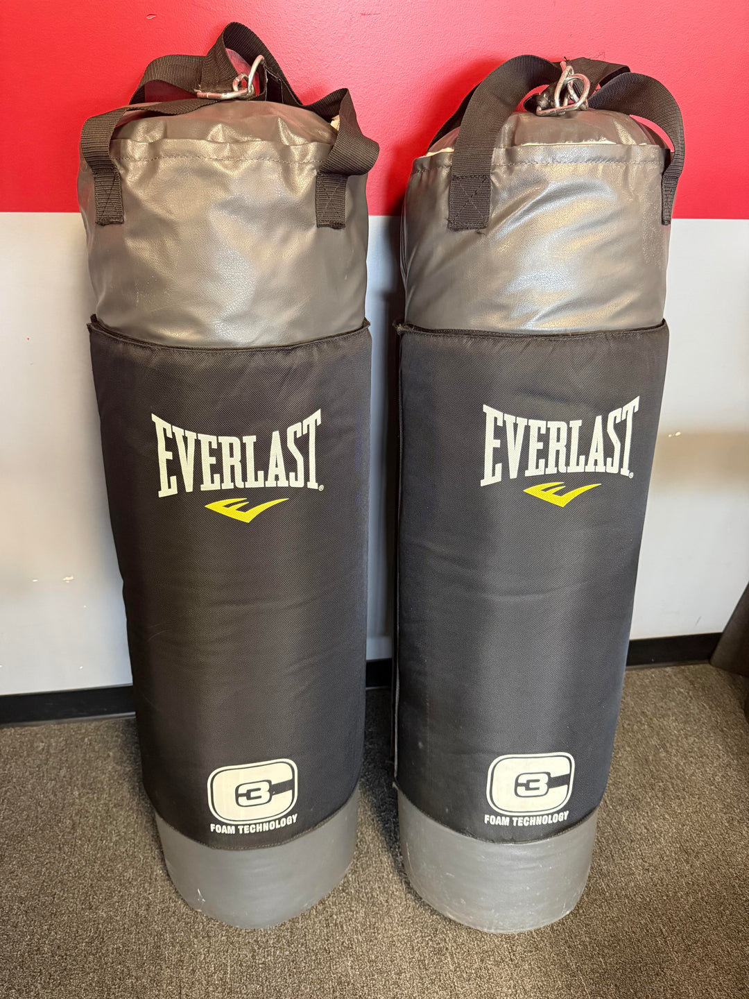 Two Everlast heavy bag punching bags standing side by side in front of a red and white wall on a carpeted floor.