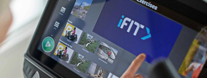 A close-up of a touchscreen exercise machine showing the iFIT app interface, with a finger touching the screen and part of a person&