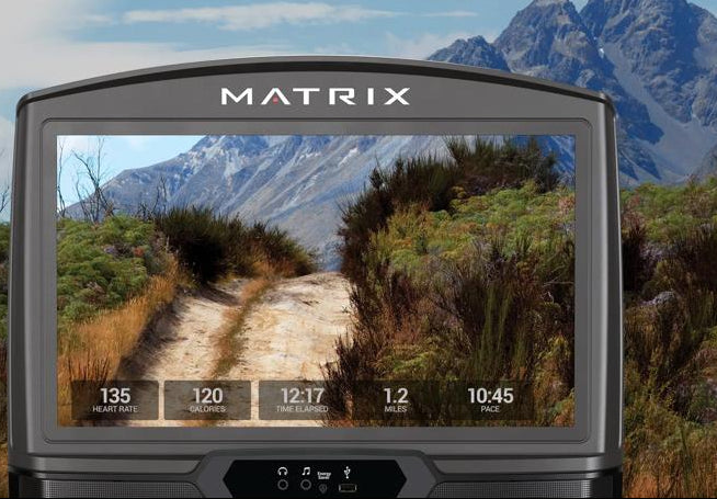 A Matrix-branded electronic device shown in the foreground with a mountain landscape displayed on its screen, set against a real outdoor mountain and brush environment. The device appears to be the product, while the outdoor scene functions as contextual background.