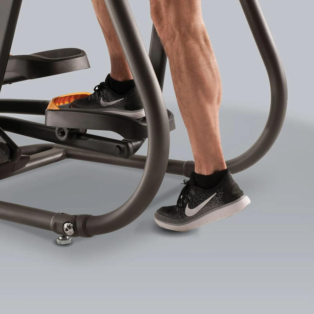 Close-up of a person using a step platform elliptical machine, showing the lower legs and feet in black Nike sneakers on the dark gray exercise equipment against a light gray studio background.