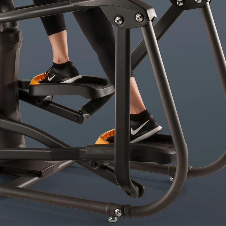 Close-up of a person using a black elliptical trainer, with their legs and feet visible stepping on the pedals against a dark blue-gray background.