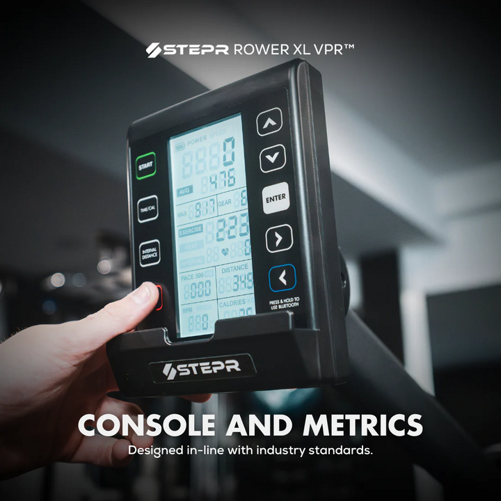 Hand holding a Stepr Rower XL VPR console with digital display and buttons, set against a blurred indoor background.