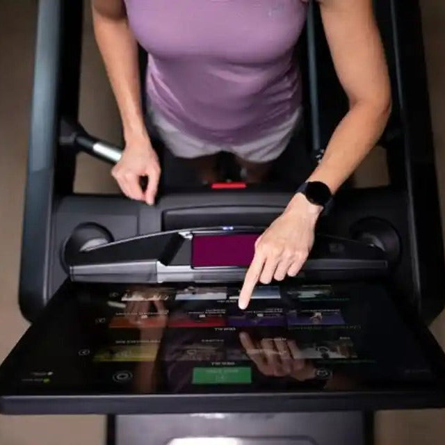 A person in a purple athletic top and white shorts walks on a treadmill while touching the touchscreen interface of the treadmill console in a gym setting.
