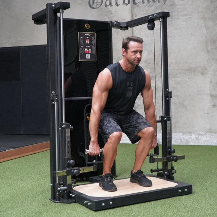 Man using a black gym machine in a room with a gray wall and green floor.
