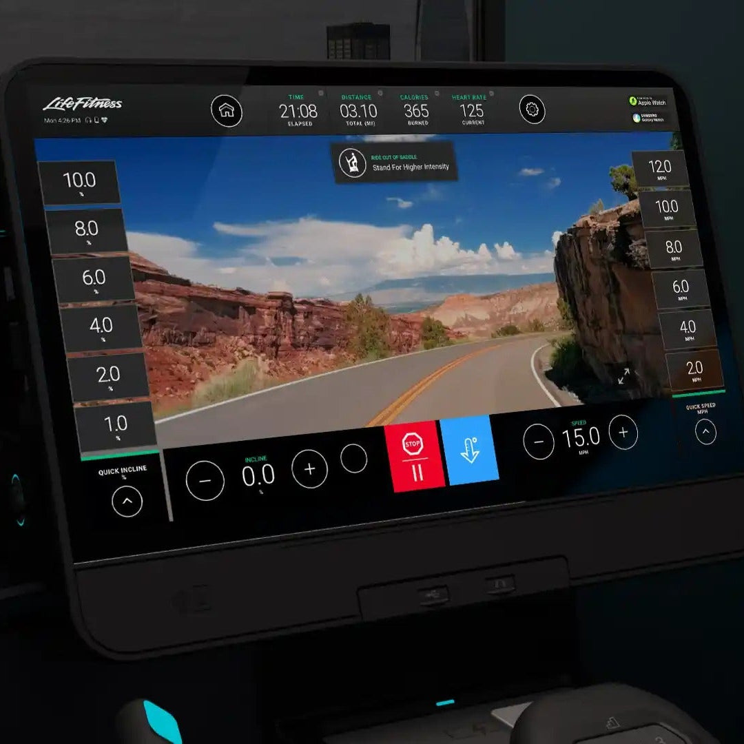 A close-up of a fitness treadmill or elliptical machine console showing a large touchscreen with a virtual outdoor road-trip scene. The device has a black frame with illuminated teal accents, and the screen displays workout metrics and controls.