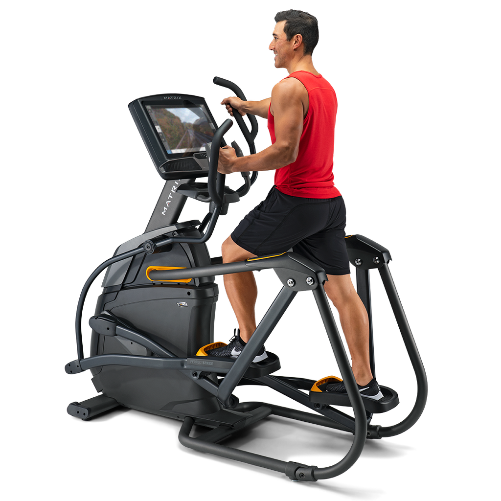 A man in a red tank top and black shorts is using a black and gray elliptical trainer with a built-in screen, shown from a side profile against a clean white background.