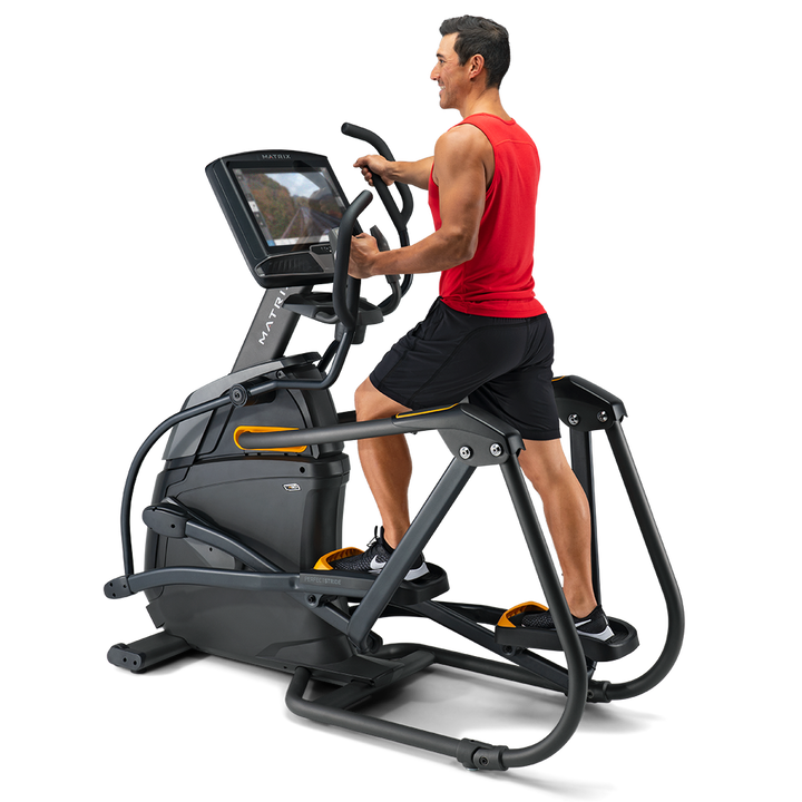 A man in a red tank top and black shorts is using a black and gray elliptical trainer with a built-in screen, shown from a side profile against a clean white background.