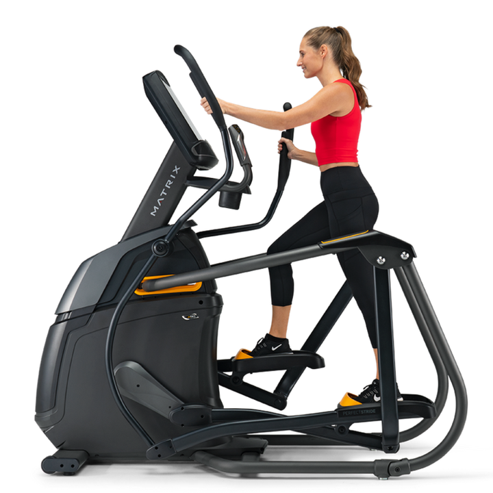 A woman in a red sports bra and black leggings is using a black and gray elliptical trainer with yellow accents on a solid white background.